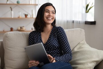 Overjoyed young Vietnamese young woman sit rest on sofa in living room look in distance using tablet, smiling millennial Asian girl relax on couch browse internet on pad gadget, have fun at home