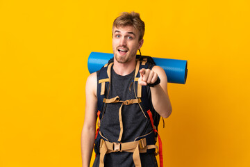 Young mountaineer man with a big backpack isolated on yellow background surprised and pointing front