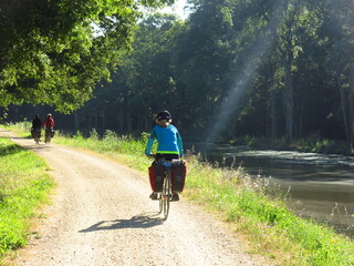 Jeune femme cycliste à vélo avec sacoche sur piste cyclable pour randonnée bicyclette le long d'un canal d'eau de rivière en France dans la Loire - obrazy, fototapety, plakaty
