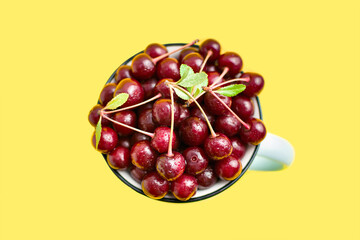 Close-up of a rustic mug filled wth fresh red ripe cherry with sprigs and water droplets in a center, top view on yellow background. Sweet fruits in a cup. Healthy seasonal food. Summertime vitamins.