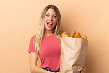 Young blonde woman holding a bag full of breads isolated on beige background with surprise facial expression