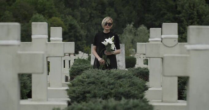 Portrait of young widow looking to camera while standing in row of stone crosses. Woman in dark glasses and clothes holding white lily flower at cemetery. Concept of memorial day