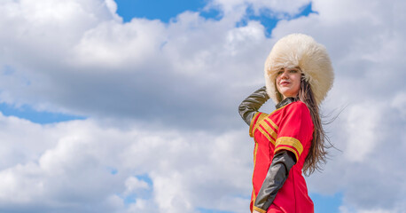 A young girl in a national Caucasian costume (Lezginka) and in a white fur hat stands against a blue cloudy sky. Summer Adventures.
