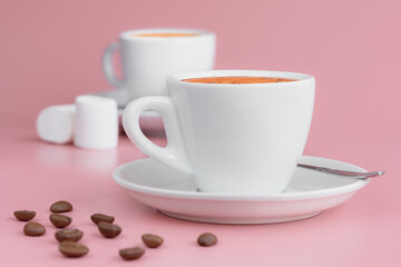 Two white cups of hot aroma coffee, some marshmallows and coffee beans on pink background. Morning coffee concept.