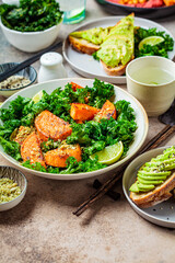 Vegan lunch table. Baked sweet potato salad with kale, avocado toasts and hummus on dark background. Plant based food concept.