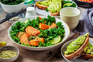 Vegan lunch table. Baked sweet potato salad with kale, avocado toasts and hummus on dark background. Plant based food concept.