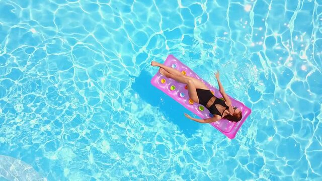 Fitted pretty girl in bikini chilling on inflatable pink mattress in swimming pool. Shaped woman in swimsuit tanning. Female relaxing on float in blue water at luxury resort. Aerial, view from above.