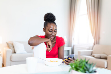 Fototapeta na wymiar Charming young african american woman sitting in the morning at the table browsing smart phone, freelancer have breakfast eat cereal corn flakes, stay home distance remote work quarantine indoors