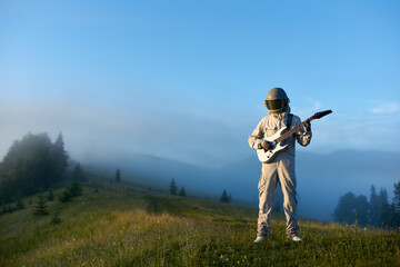 Spaceman in white space suit and helmet playing white guitar, standing on sunny green mountain glade in the morning, foggy hills and blue sky on background. Concept of astronautics, music and nature.
