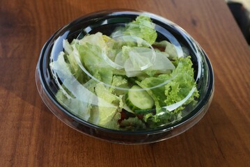 salad of fresh green vegetables in a closed transparent plastic bowl on a brown table