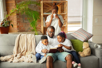 Obraz premium Reading. Young and cheerful african family during quarantine, insulation spending time together at home. Time for family, happiness and love. Concept of quarantine lifestyle, togetherness, home