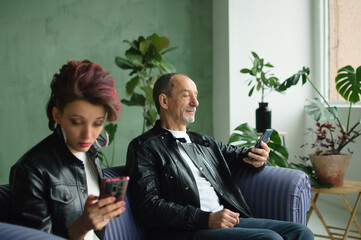 Family portrait of adult daughter and senior father in loft room with houseplants. Man and girl are wearing black leather jackets in punk style and browsing on their smartphones without communication
