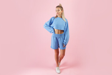 Fashionable sportswear look on young caucasian blonde woman. Blue shorts and sweatshirt. Isolated on pink background