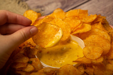 The girl takes a chip from a round dish with potato chips and a saucepan with cheese sauce in the center of the plate. Close up.