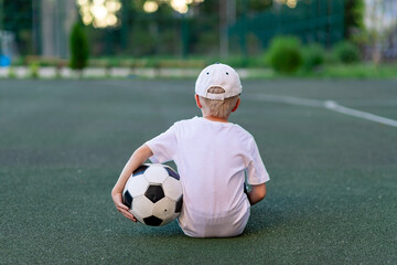 a boy in sports clothes sitting on a green lawn on a football field with a soccer ball back, back view, sports section, training