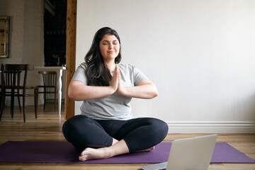 Overweight barefoot young female in sportswear sitting on yoga mat with eyes closed, meditating,...