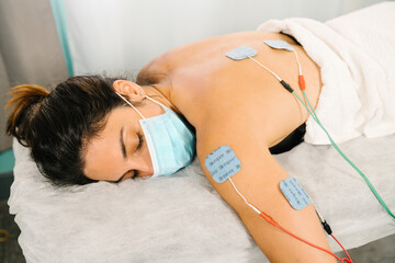 Caucasian woman receiving physiotherapy treatment with electro-stimulation electrodes on her back lying on a stretcher with a face mask