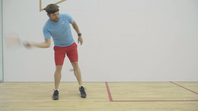 Wide shot of strong Caucasian mid-adult sportsman training in gym. Portrait of brunette bearded athletic man exercising indoors. Confident concentrated guy playing squash racket and ball sport.