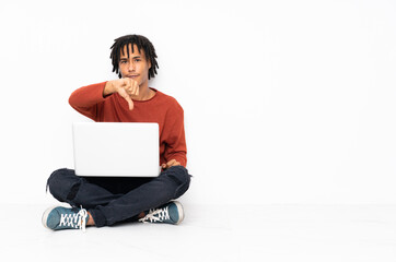 Young african american man sitting on the floor and working with his laptop showing thumb down with...