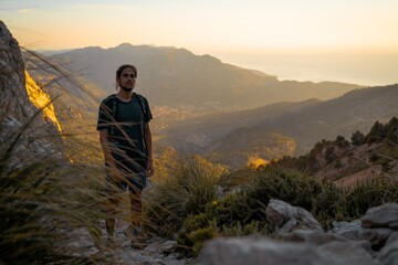 Young spanish man standing on a rock looking at camera while hiking in Serra de Tramuntana at golden hour with the village of Soller at the backgorund tinted by golden light