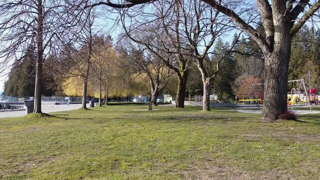 Ground hold shot. Grass, trees, playground near seawall. People walking. Taped off playground. Closed due to physical distancing. Outdoor pool in background.