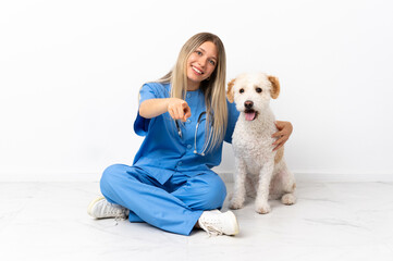 Young veterinarian woman with dog sitting on the floor pointing front with happy expression