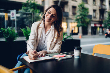 Cheerful hipster girl smiling while doing test tasks enjoying pastime in sidewalk cafeteria, happy Caucasian female writer creating publication text sitting at street table with textbook