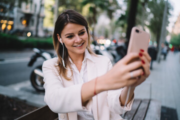 Pretty cheerful girl in earphones posing for selfie making via smartphone camera and listening favourite playlist, female hipster taking picture enjoying music via cellular player during leisure