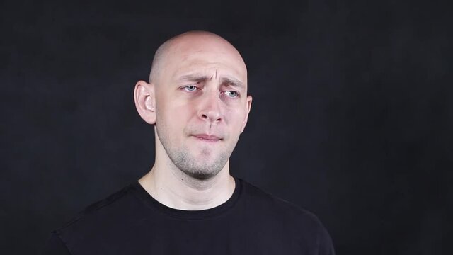 A bald caucasian man in a black t-shirt on a black background depicts a grimace of discontent close-up. High quality FullHD footage