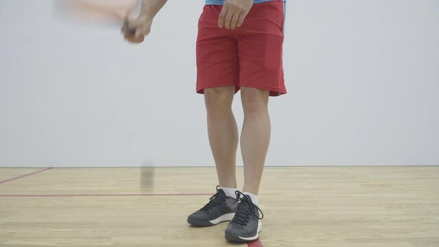 Unrecognizable Caucasian sportsman in sport shorts and sneakers hitting squash ball with racket against the floor. Athletic man practicing sport game in gym indoors.