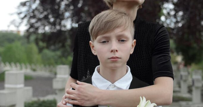 Upset boy holding flower and looking to camera while his mom embracing him. Young widow in dark clothes hugging her teen son while they standing at cemetery. Concept of memorial day