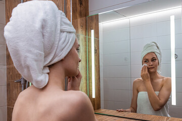 Young woman after a shower standing in the bathroom in front of the mirror with a towel wrapped on her head and washes her face with a cosmetic cotton pad with lotion. Skin care