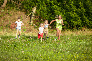 Naklejka na ściany i meble Kids, children running on meadow in summer's sunlight. Look happy, cheerful with sincere bright emotions. Cute caucasian boys and girls. Concept of childhood, happiness, movement, family and summer.
