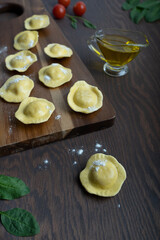 Uncooked italian ravioli pasta on cutting board surrounded by olive oil gravy boat, spinach leaves and tomatoes on dark brown wooden background. Image with selective focus, vertical orientation