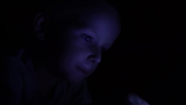 The little boy lies in bed before going to bed and plays on the smartphone. Night shot.