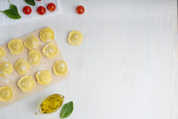 Top view of raw italian ravioli pasta on cutting board surrounded by olive oil gravy boat, spinach and tomato on towel on white wooden background. Image with copy space, horizontal orientation