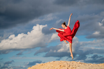young ballerina in a bright red long dress soars in a jump above the ground, like a bird, against a...