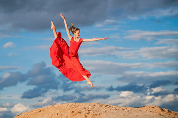 young ballerina in a bright red long dress soars in a jump above the ground, like a bird, against a background of blue sky and clouds. The skirt develops like wings. © Maria Moroz
