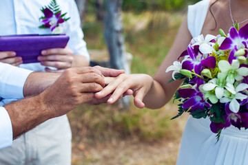 Fototapeta premium close-up of the groom puts the ring on the bride's finger. The presenter of the ceremony is out of focus in the background. White and purple colors. Outdoors