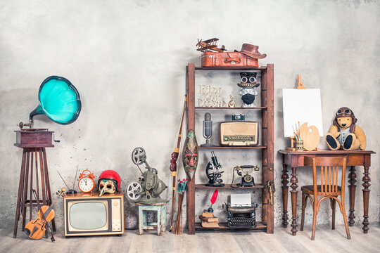 Collection of antique media devices, writers tools, gramophone, film projector, old Teddy Bear toy with canvas blank on easel, travel bag front concrete wall background. Vintage style filtered photo