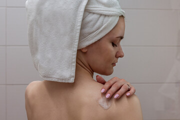 Beautiful young woman with a white towel on her head standing in the bathroom and applying shower foam to her shoulder.