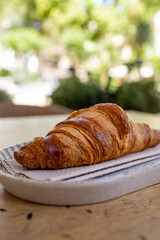 Summer breakfast in vacaton. A beautiful plain french butter croissant on a plat on the outdoor table of a bar in a touristic destination.