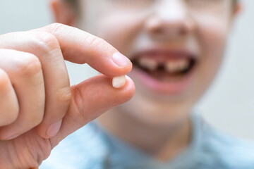 The child's hand shows the fallen out small white milk tooth close-up and smiles with a hole in the...