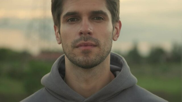 A close-up view of a young sportsman in hoodie is opening his eyes and looking to the camera while walking outside