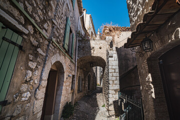 A cobbled street with a stone arch and dry grass shows of how it must have looked like in a medieval village. Narrow alleyway with tall stone walls in the old town - St Paul de Vence, France