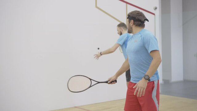 Two positive concentrated sportsmen playing squash in gym. Wide shot of confident Caucasian men training indoors. Smiling bearded guys practicing sport game in sports club.