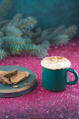 Obraz na płótnie Canvas A green mug of cocoa with cream and pieces of chocolate on a background of fir branches, on a bright pink table. Christmas mood