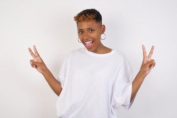 Indoor portrait of young caucasian female isolated on gray background with optimistic smile, showing peace or victory gesture with both hands, looking friendly. V sign.