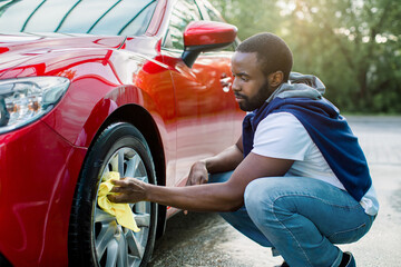 Car washing on open air. Young hipster African bearded man cleaning a wheel, car rims of modern luxury red car with yellow microfiber cloth, at outdoor car wash self service