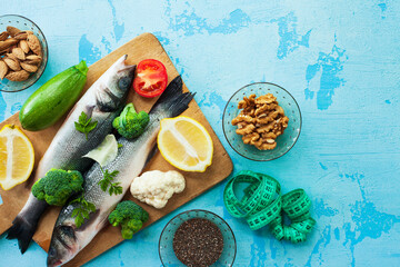 Measuring tape, fresh raw fish sea bass on cutting board, raw vegetables, spices and nuts. Stone background. Healthy food, lifestyle, diet and weight control concept. Space for a text.